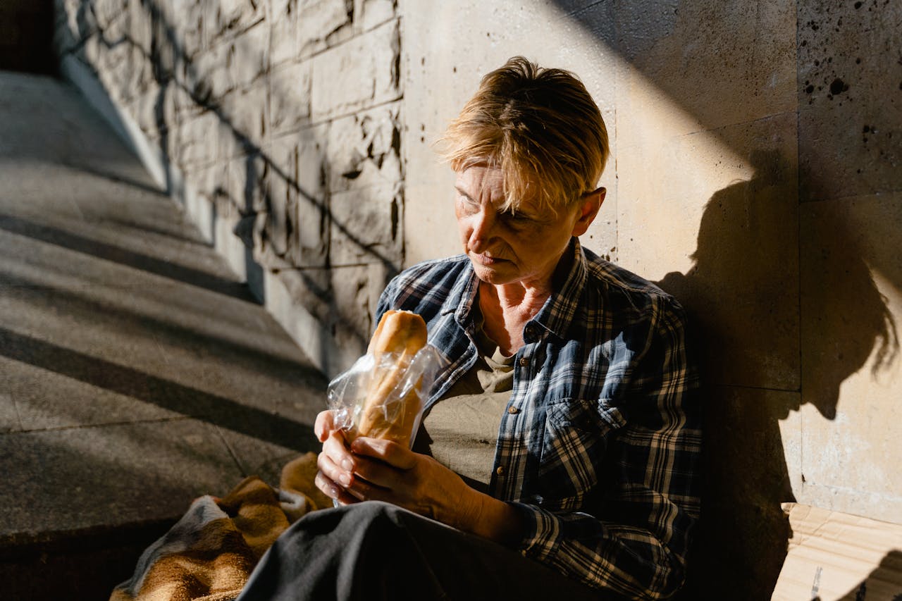 services-04 A senior woman holding and eating bread while seated against a sunlit wall, casting a thoughtful shadow.